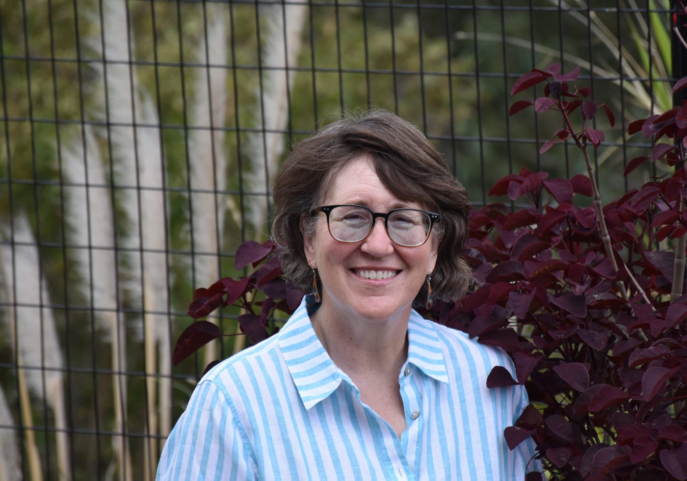 Lynne Klippel smiling in a blue striped shirt beside a garden fence